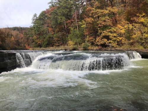 A serene waterfall cascades over rocks, surrounded by vibrant autumn foliage and a calm river.