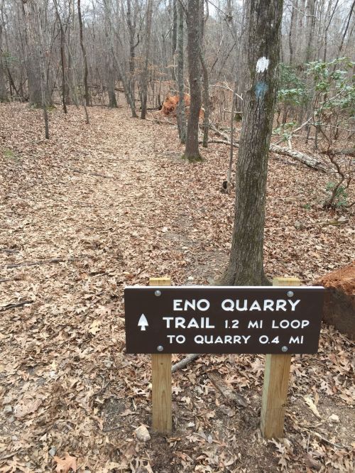 A wooden sign for Eno Quarry Trail, indicating a 1.2-mile loop and a 0.4-mile path to the quarry, surrounded by trees.