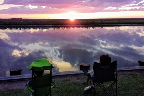 Two children sit in lawn chairs by a calm river, watching a vibrant sunset reflected in the water.