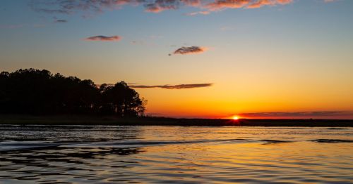 A serene sunset over calm waters, with silhouettes of trees against a colorful sky.