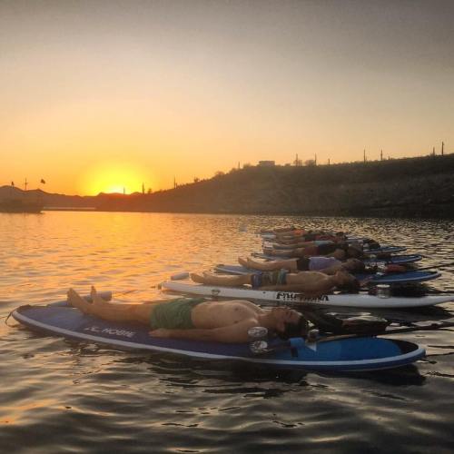 A group of people lying on paddleboards at sunset, surrounded by calm water and a scenic landscape.