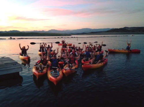 A group of people in kayaks cheer and raise their paddles on a lake at sunset, with mountains in the background.