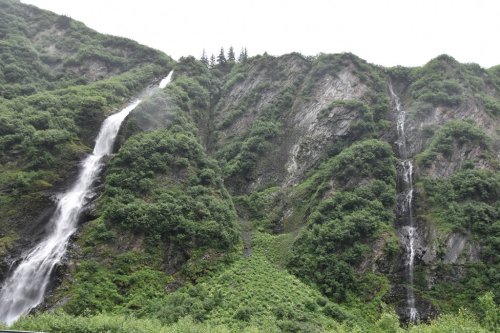 Two waterfalls cascade down a lush, green mountainside, surrounded by dense vegetation and rocky terrain.