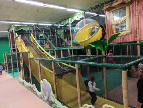 Colorful indoor playground with slides, climbing structures, and a large yellow ball, filled with children playing.