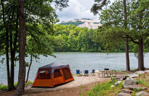 A tent by a serene lake, surrounded by trees, with camping chairs and a picnic table nearby, under a cloudy sky.