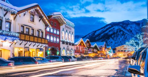 A snowy street scene with festive lights, charming buildings, and mountains in the background at dusk.