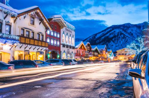 A snowy street lined with festive buildings and lights, with mountains in the background and car lights creating streaks.