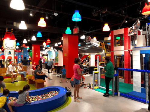 A colorful indoor play area with children playing, adults chatting, and various activity zones featuring toys and structures.