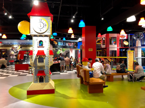 Colorful indoor play area featuring a large LEGO clock tower, seating, and families enjoying the space.