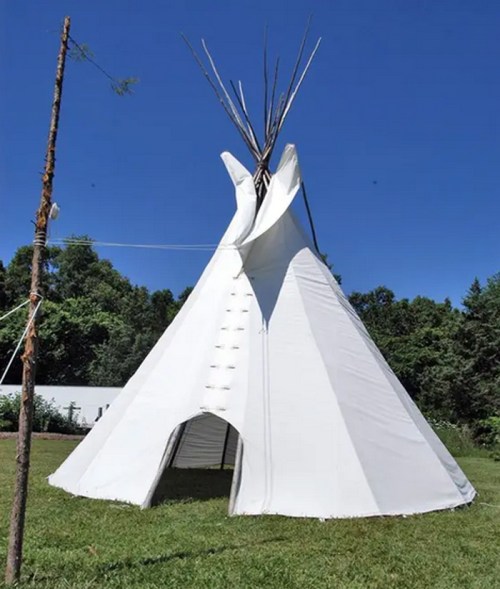 A white teepee tent stands on green grass under a clear blue sky, with wooden poles at the top.