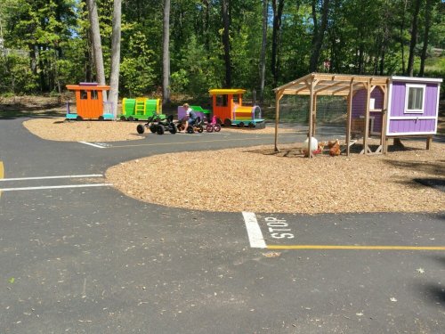 Colorful play structures and a chicken coop in a gravel area surrounded by trees, with a child riding a toy vehicle.