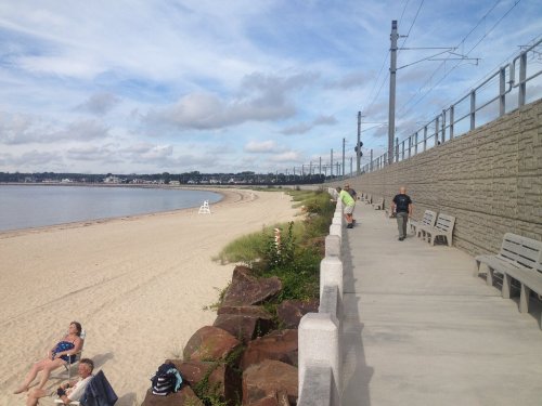 A sandy beach with a walkway, benches, and people enjoying the sun, alongside a train track and cloudy sky.
