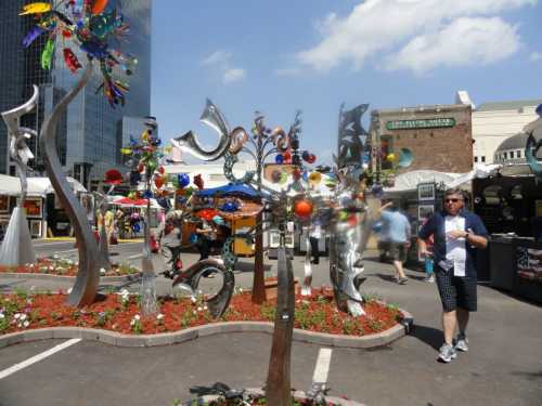 A vibrant outdoor art display featuring colorful sculptures and flowers, with a person walking nearby.