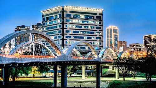 A modern bridge with arches spans a river, with a city skyline illuminated at dusk in the background.