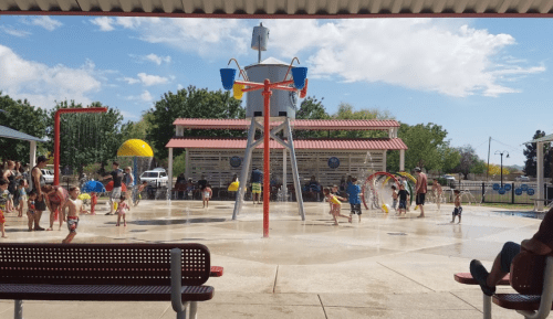A splash pad with children playing in water, surrounded by trees and a sunny sky. Benches in the foreground.