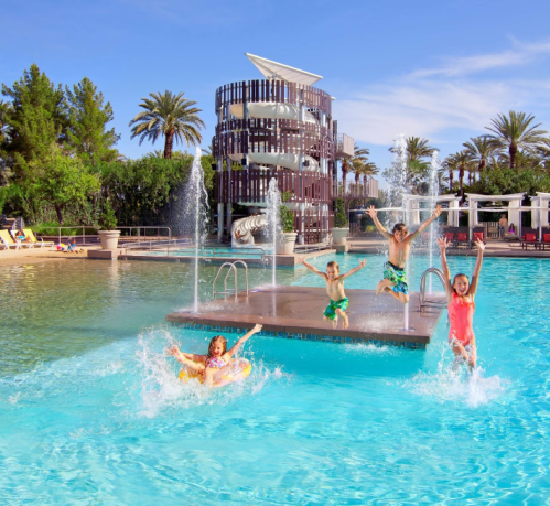 Children joyfully jumping into a sparkling pool with fountains, surrounded by palm trees and a modern building.