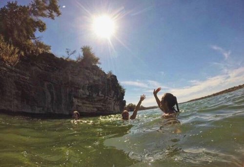 Three people swimming in clear water near a rocky shore, with a bright sun shining overhead.