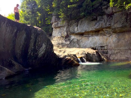 A person stands on a rocky ledge above a clear, green pool surrounded by trees and cliffs.