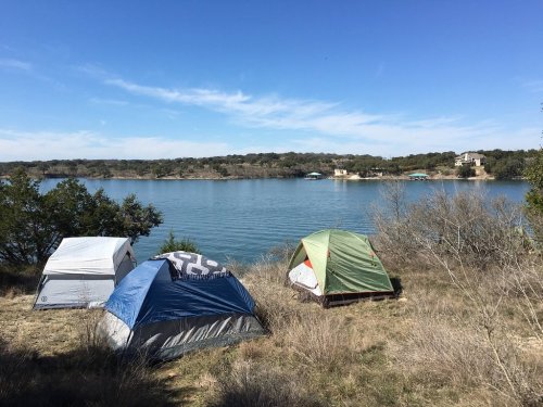Three tents set up by a calm lake, surrounded by grass and trees under a clear blue sky.