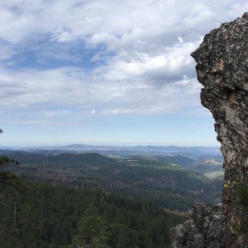 A rocky outcrop overlooks a vast green valley and distant mountains under a blue sky with scattered clouds.