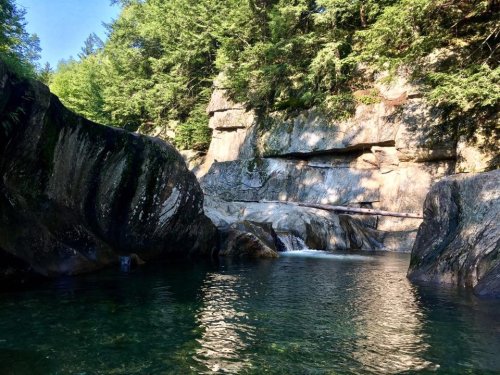 A serene natural scene featuring a clear pool of water surrounded by large rocks and lush green trees.