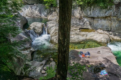 A serene river scene with a waterfall, rocky banks, and colorful towels laid out on the smooth stones.