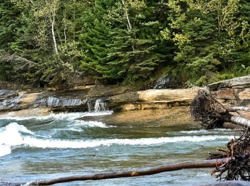 A serene river flows past rocky banks and lush green trees, with gentle waves and fallen branches along the shore.