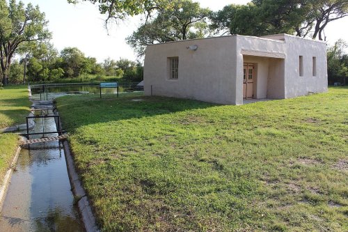 A small, light-colored building beside a grassy area and a narrow water channel under a clear sky.