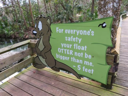 A cartoon otter on a sign warns floaters to stay within a 5-foot limit for safety, set in a natural park setting.