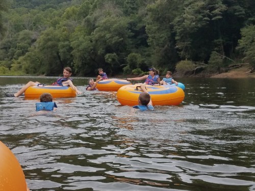 A group of children and teens float on orange inner tubes in a river surrounded by trees.