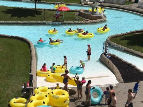 A water park scene with people floating on yellow inner tubes in a lazy river, surrounded by green grass and umbrellas.