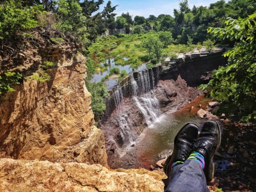 A person's feet in black shoes with colorful socks resting on a rocky ledge, overlooking a waterfall and lush greenery.