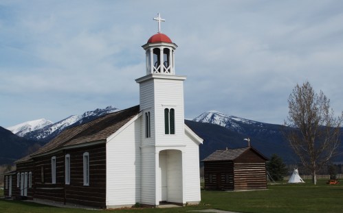 A white church with a red dome and cross, set against a backdrop of snow-capped mountains and a clear sky.