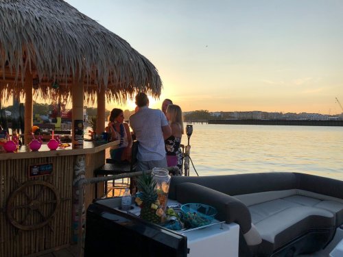 A group of people socializing at a tiki bar by the water during sunset, with a cozy seating area nearby.