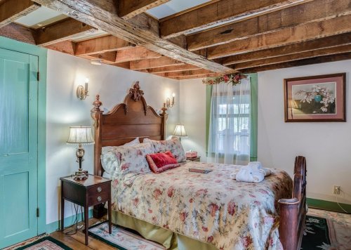 Cozy bedroom with a floral bedspread, wooden beams, and soft lighting, featuring a nightstand and decorative artwork.
