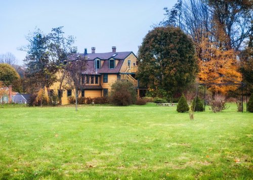 A large yellow house surrounded by trees and a grassy lawn, with autumn foliage in the background.