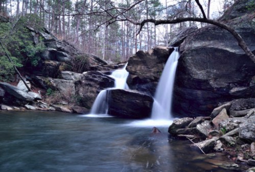A serene waterfall cascades over rocks into a calm pool, surrounded by lush greenery and trees.