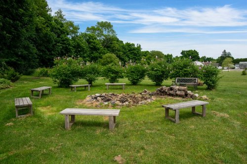 A circular fire pit surrounded by wooden benches in a grassy area with trees and a blue sky in the background.