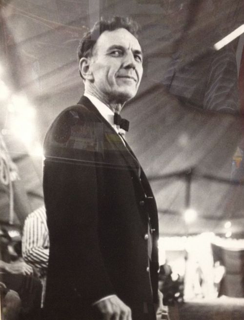 A black and white portrait of a man in a tuxedo, looking confidently to the side, with a tent backdrop.