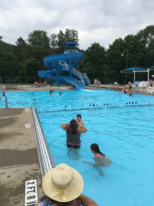 A busy outdoor pool with a water slide, people swimming, and lounging on deck chairs under a cloudy sky.