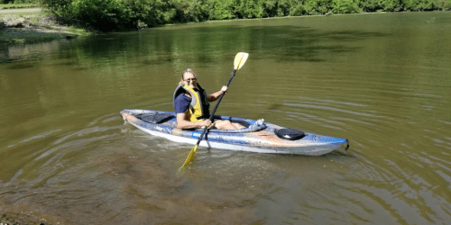 A person in a kayak paddles on a calm river, surrounded by greenery and sunlight.