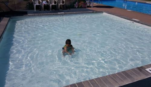 A child sits in a shallow pool, surrounded by clear water and poolside chairs in the background.