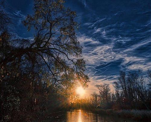 A serene sunset over a calm river, framed by silhouetted trees and a dramatic sky filled with clouds.