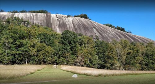 A large, smooth rock formation rises above a grassy field, surrounded by trees under a clear blue sky.