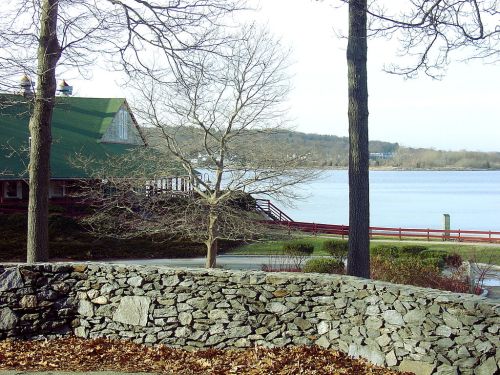 A serene lakeside view featuring a stone wall, bare trees, and a green-roofed building by the water.