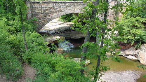 A stone arch bridge over a small waterfall, surrounded by lush greenery and rocky terrain.