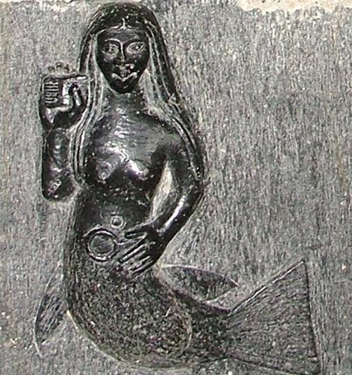 A carved figure of a mermaid holding a small object, with long hair and a fish tail, set against a textured background.