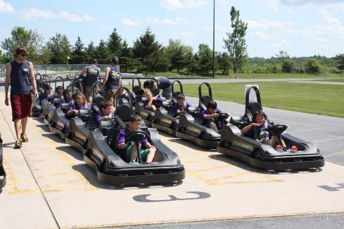 A group of children in go-karts lined up on a track, with an adult supervising them in a sunny outdoor setting.