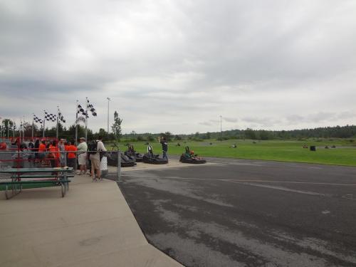 A go-kart track with racers preparing to race, surrounded by spectators and flags in a cloudy sky.