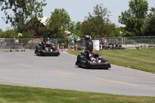 Two go-karts racing on a paved track, surrounded by greenery and spectators in the background.
